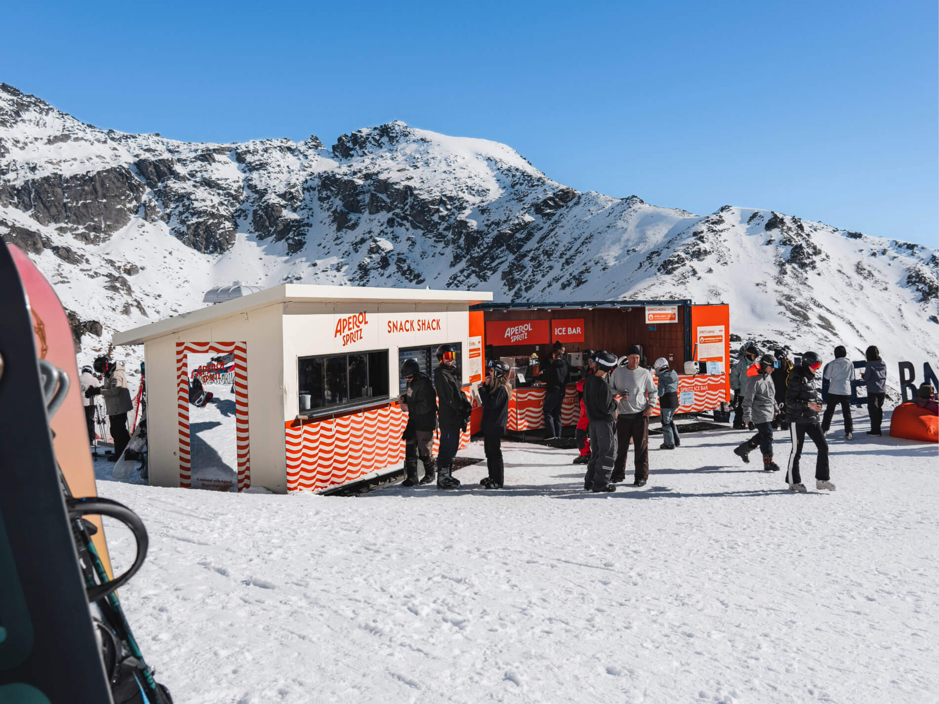 People in winter gear near Aperol Spritz snack shack and ice bar on a snowy mountain under blue sky.