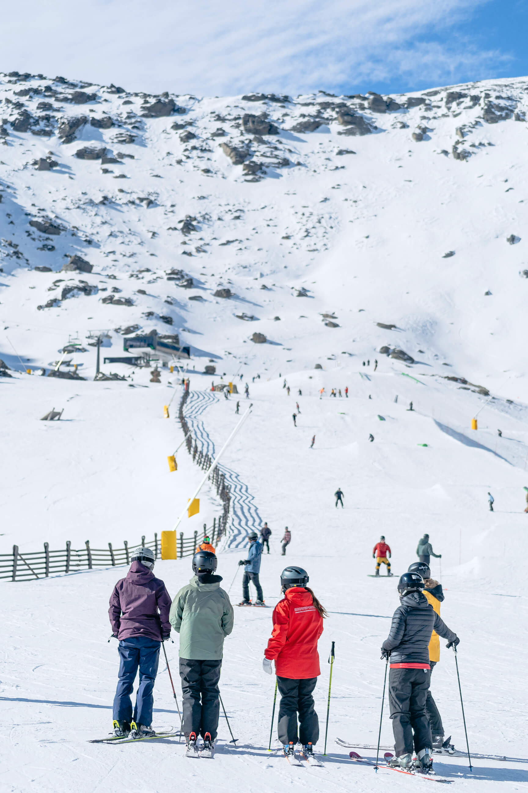 Group of skiers on a snowy mountain slope with rocky peaks and a clear sky above.